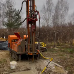 Pomarańczowa wiertnica na gąsienicach wierci w gruncie; wokół błoto i białe rury; krajobraz wiejski w tle.