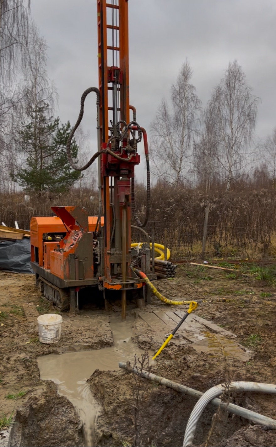 Pomarańczowa wiertnica na gąsienicach wierci w gruncie; wokół błoto i białe rury; krajobraz wiejski w tle.