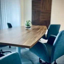 Sala konferencyjna z drewnianym stołem, szarymi krzesłami na kółkach i rośliną w szklanym wazonie na stole. W tle drewniana dekoracja ścienna w kwadraty i zasłonięte okno.