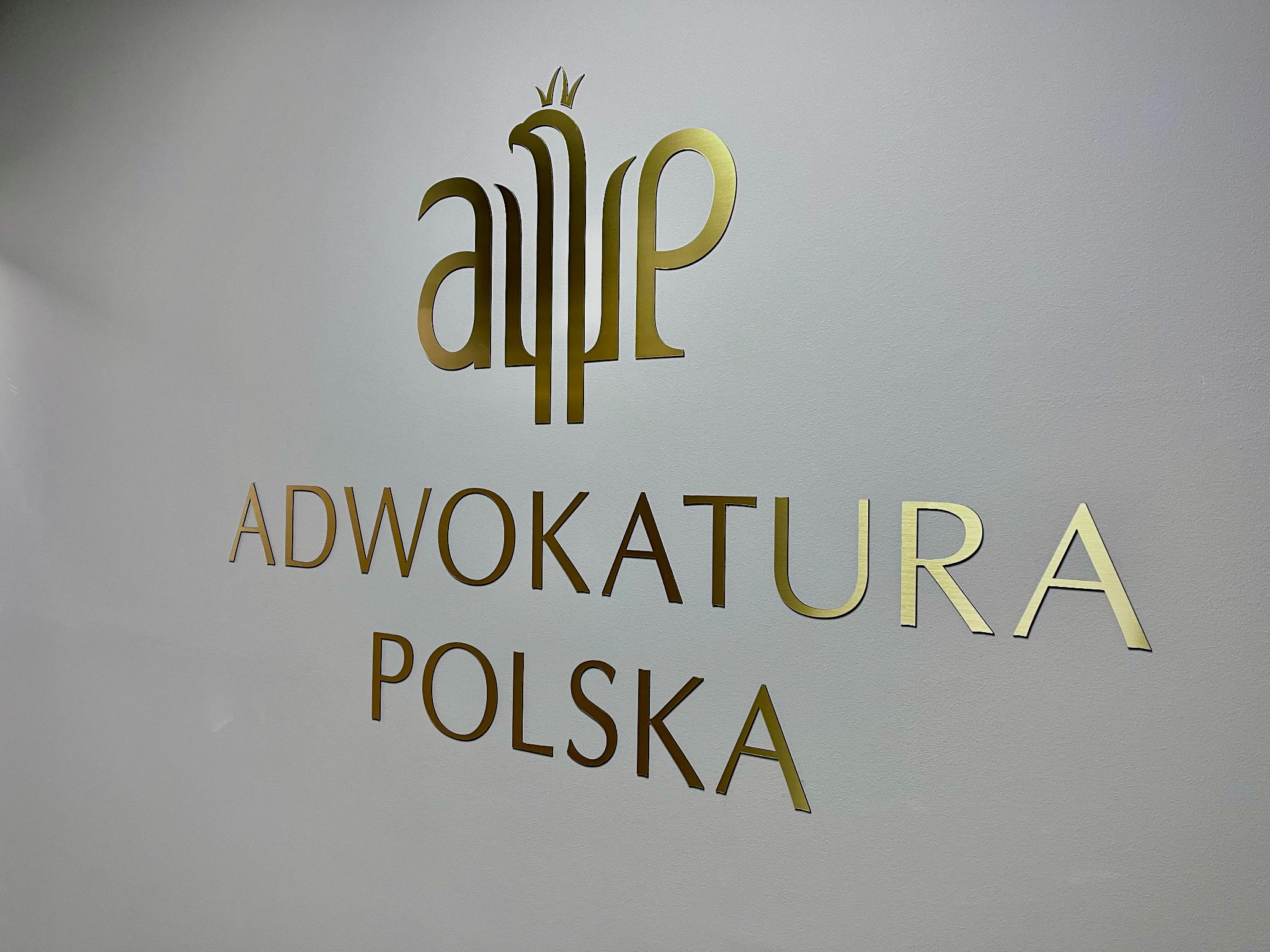 Złoty napis 'Adwokatura Polska' z stylizowanym logo na białej ścianie, oświetlenie górne.
