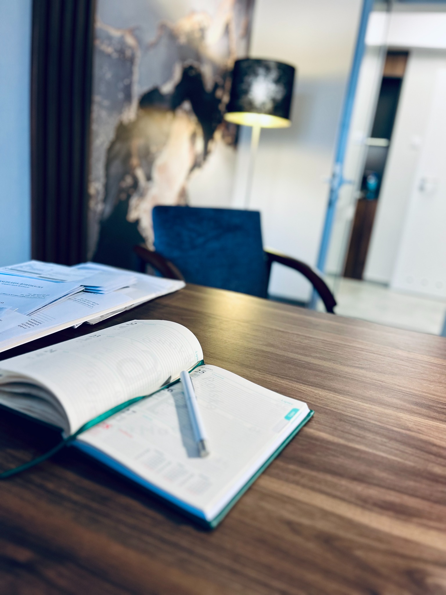 Otwarty kalendarz z długopisem na drewnianym biurku w kancelarii, w tle dokumenty, krzesło i lampa podłogowa.