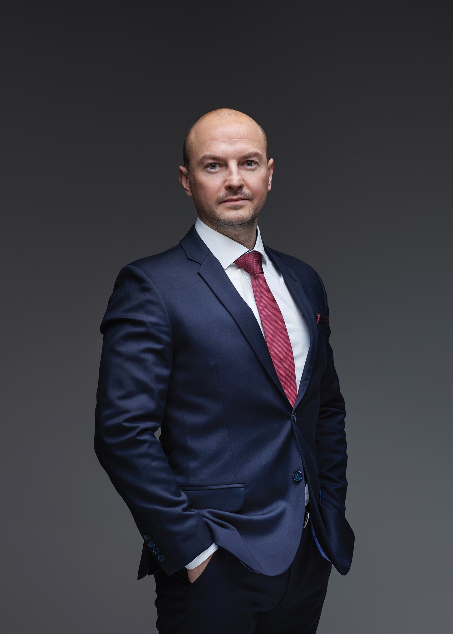 Portret mężczyzny w granatowym garniturze i czerwonym krawacie, z rękami w kieszeniach, na szarym tle.