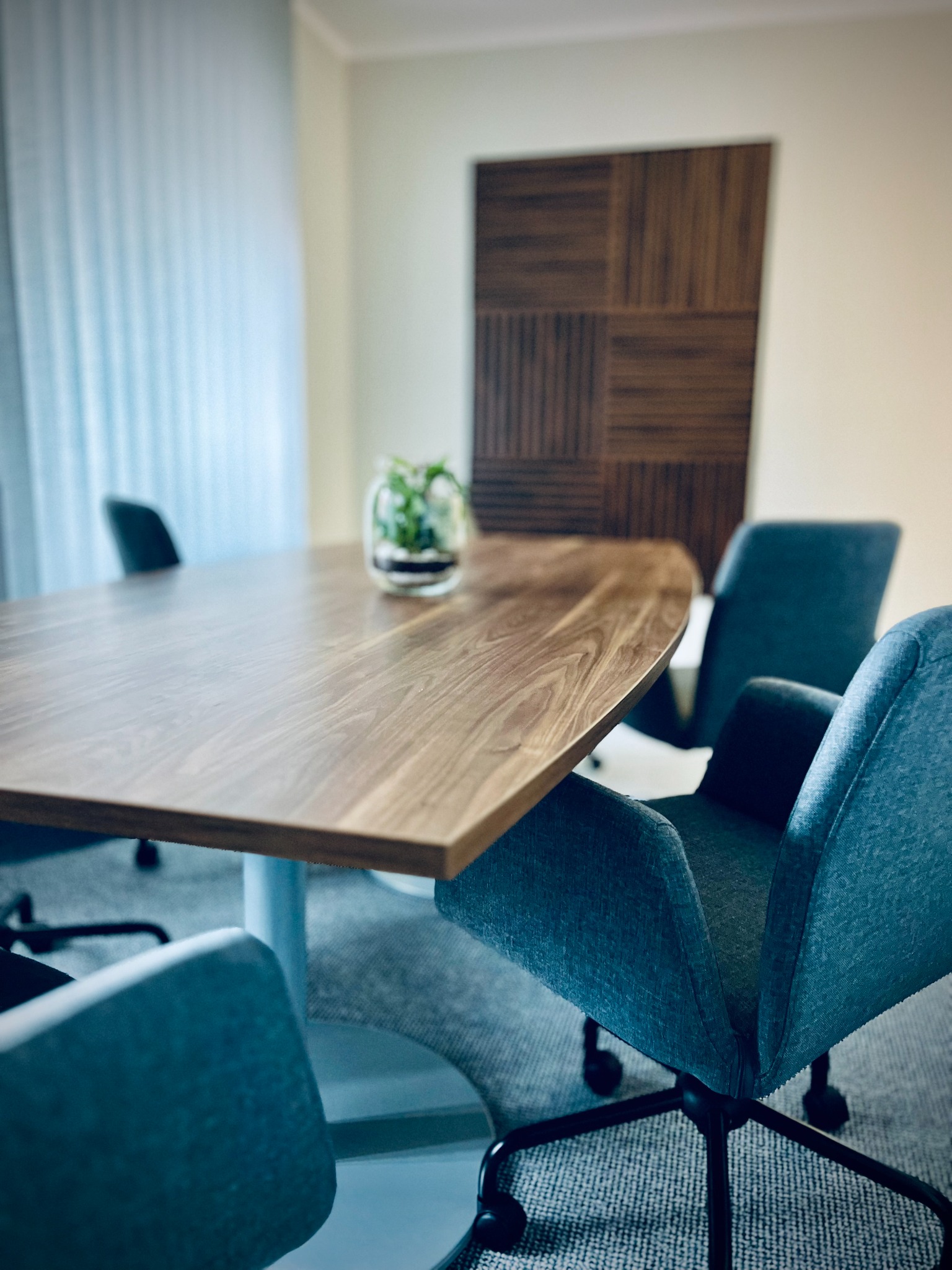 Sala konferencyjna z drewnianym stołem, szarymi krzesłami na kółkach i rośliną w szklanym wazonie na stole. W tle drewniana dekoracja ścienna w kwadraty i zasłonięte okno.
