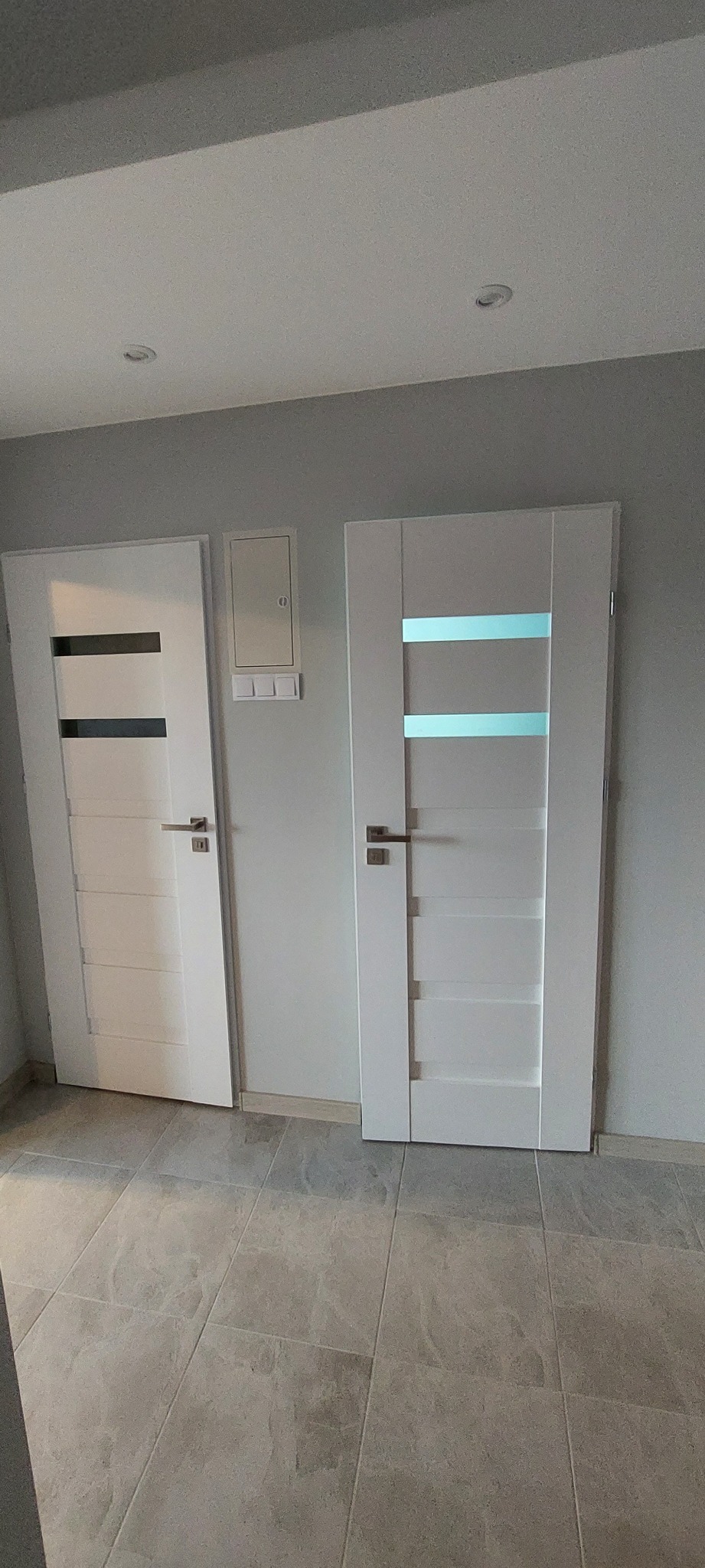 Dwa białe, nowoczesne skrzydła drzwiowe z przeszkleniami, zamontowane w szarej ścianie, widoczne z perspektywy podłogi wyłożonej płytkami imitującymi kamień, obok skrzynka elektryczna.