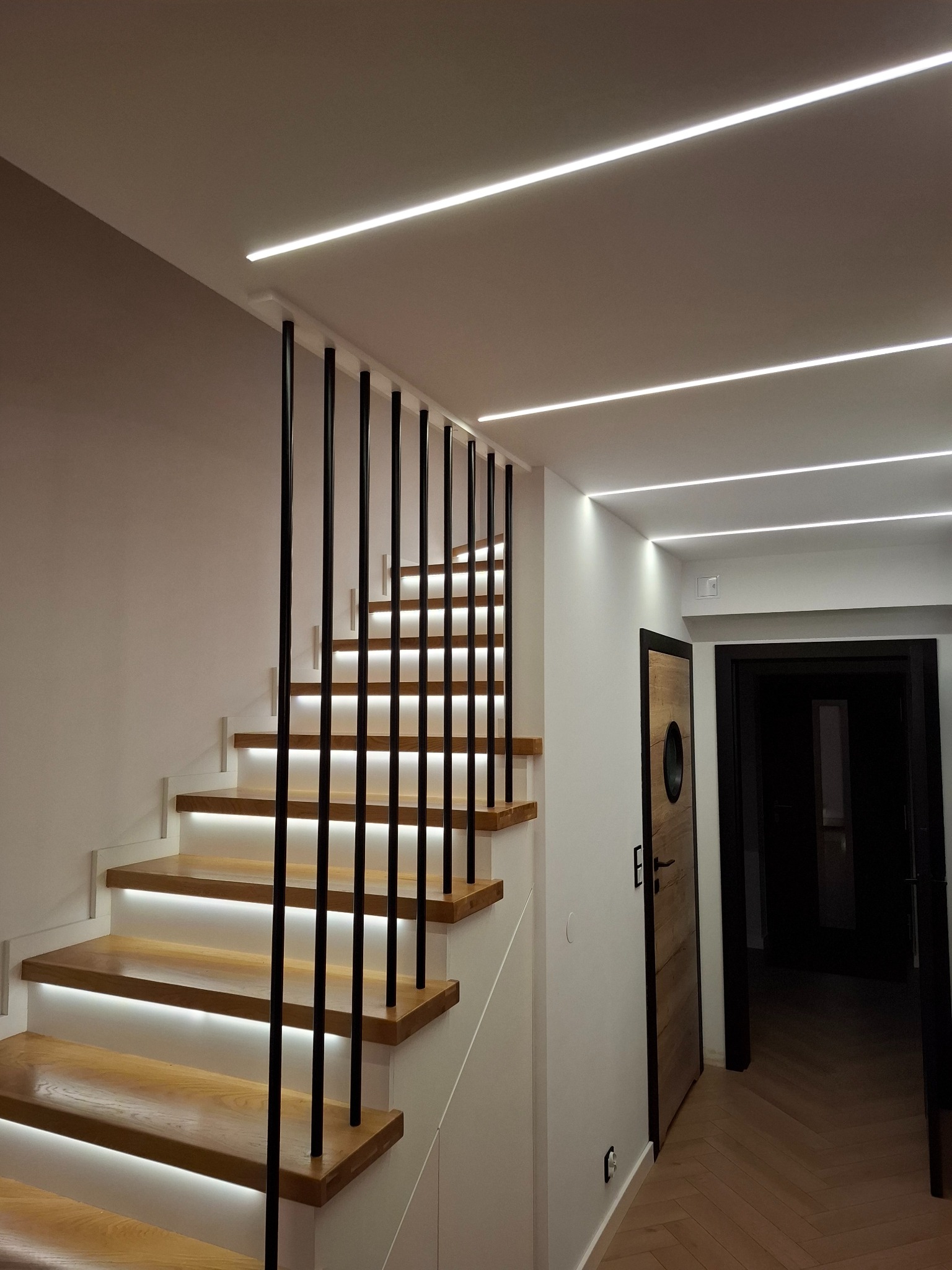 Nowoczesne schody z drewnianymi stopniami podświetlonymi ledami, czarne metalowe poręcze, sufit z liniowym oświetleniem LED, oraz drewniane drzwi z okrągłym okienkiem.