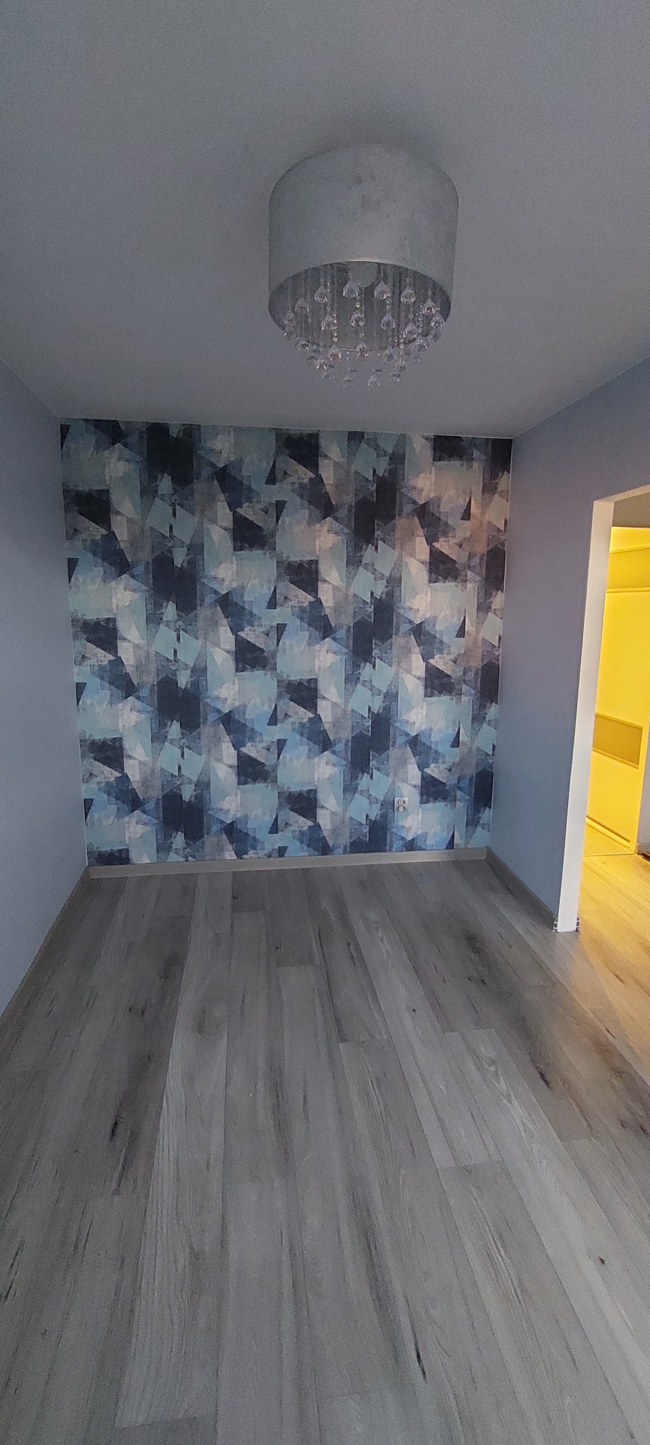 Pusty pokój po remoncie z szaro-niebieską tapetą w geometryczne wzory na jednej ścianie, szarą podłogą z paneli i nowoczesną lampą z kryształkami.