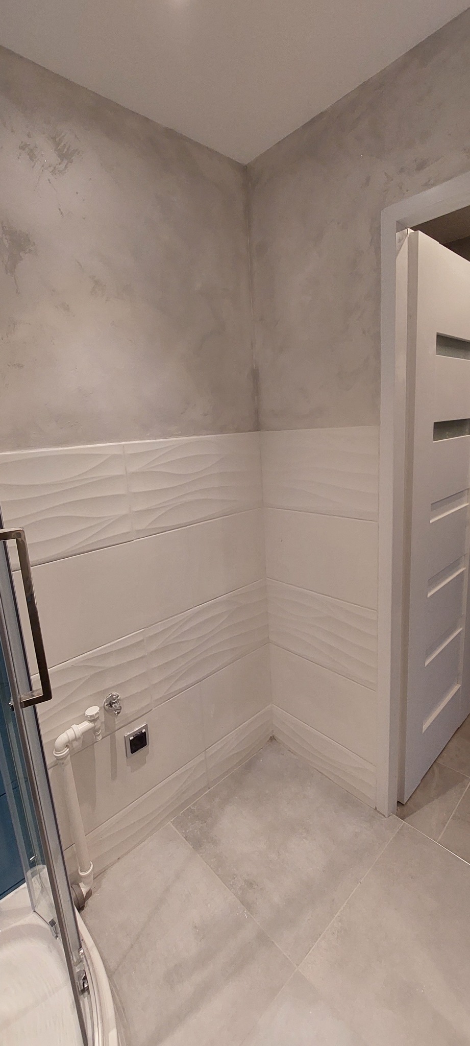 Wykończona łazienka z szarą, dekoracyjną ścianą imitującą beton, białe płytki z falistym wzorem, szara podłoga i fragment kabiny prysznicowej z chromowanym uchwytem oraz białe drzwi z poziomymi...