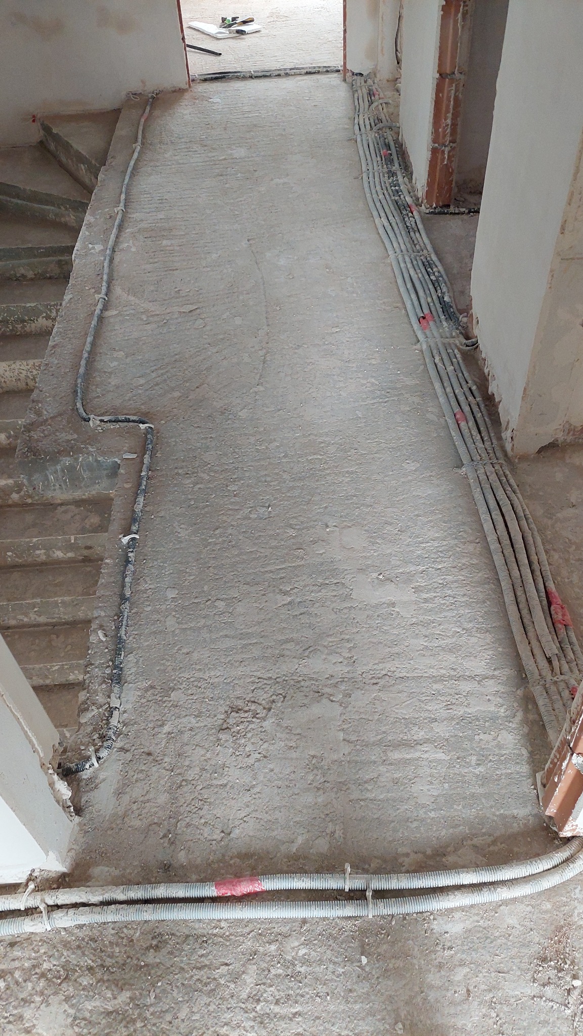 Surowy betonowy podkład podłogowy w trakcie remontu z widocznymi kablami elektrycznymi w peszlach ułożonymi wzdłuż ściany i progów drzwiowych.