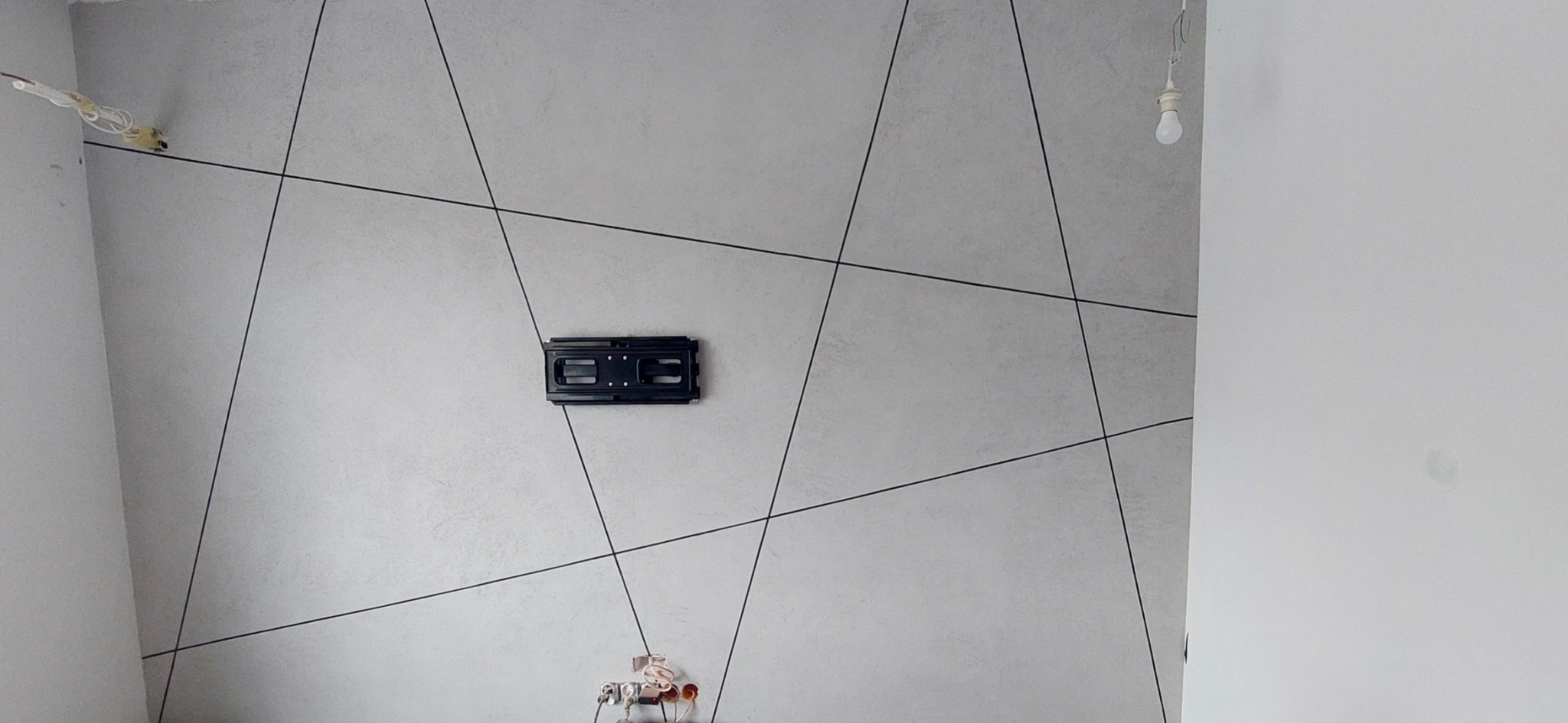 Szpachlowana ściana w jasnych odcieniach szarości z dekoracyjnymi, czarnymi liniami, uchwyt ścienny do telewizora na środku, widoczne gniazdka elektryczne i wisząca żarówka.