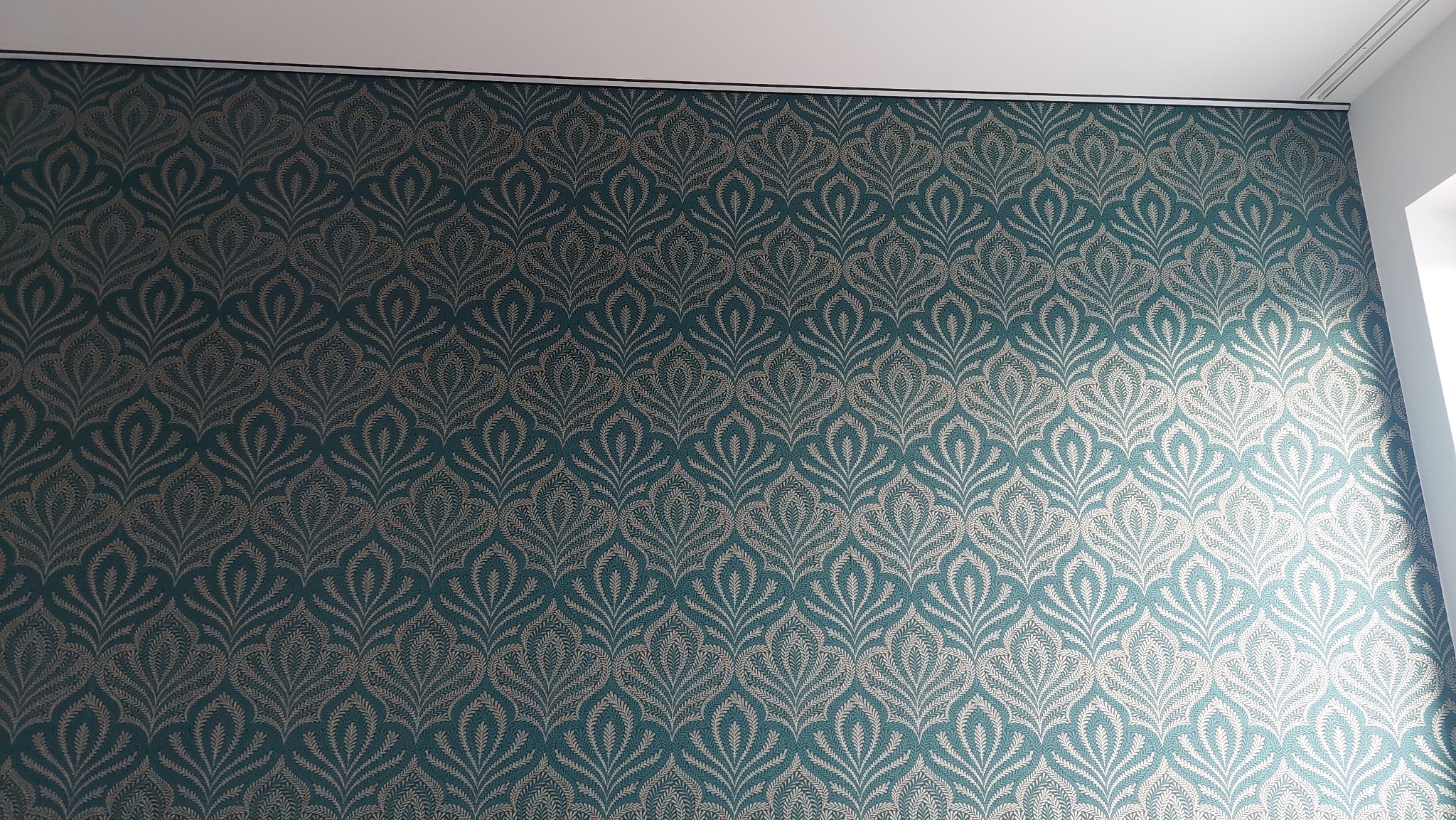 Ściana pokryta wzorzystą tapetą w odcieniach turkusu i złota, z motywem stylizowanych liści, widoczna od góry do dołu, z fragmentem sufitu i rogu ściany.