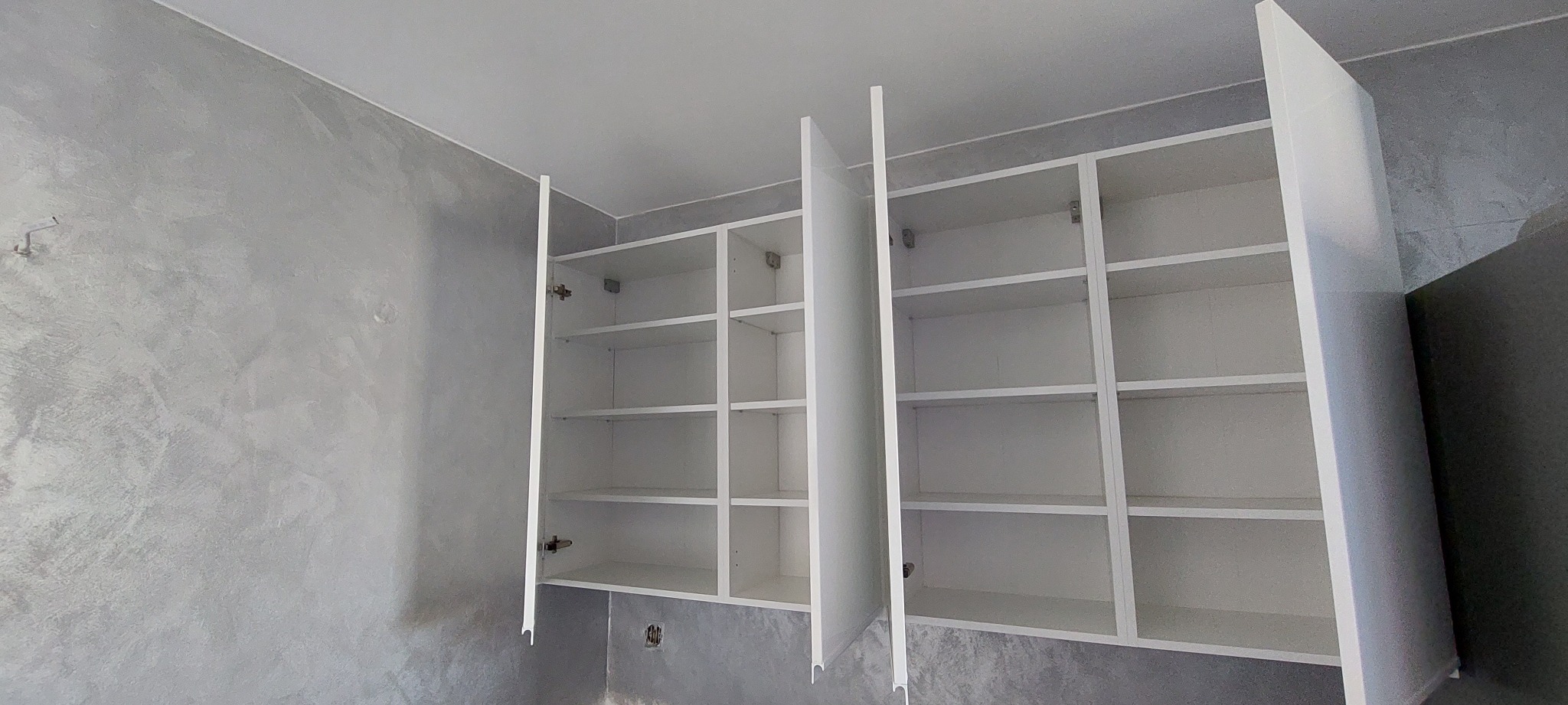 Białe, puste szafki kuchenne pod sufitem, z otwartymi drzwiczkami, na tle ściany z szarą, dekoracyjną fakturą.