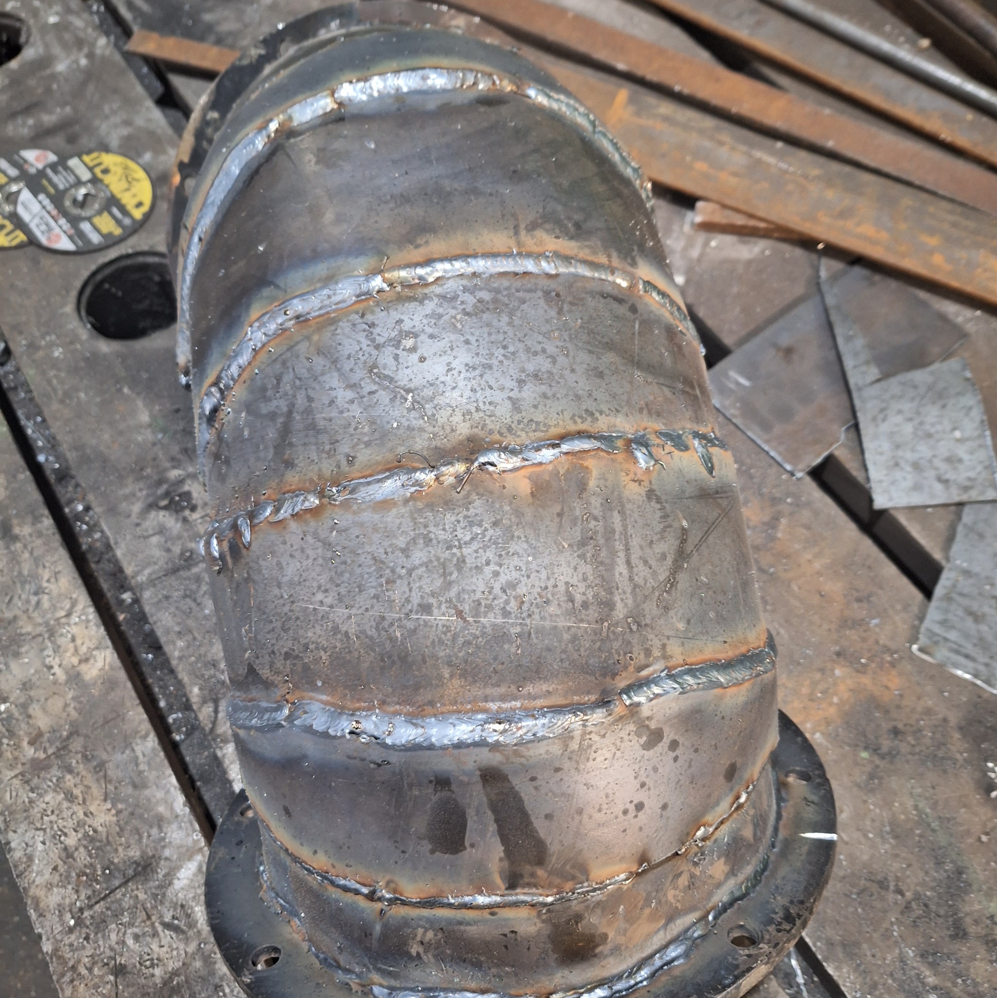 Spawany element metalowy z widocznymi spawami i kołnierzem, leżący na metalowym stole warsztatowym, obok leżą tarcze tnące i metalowe pręty.