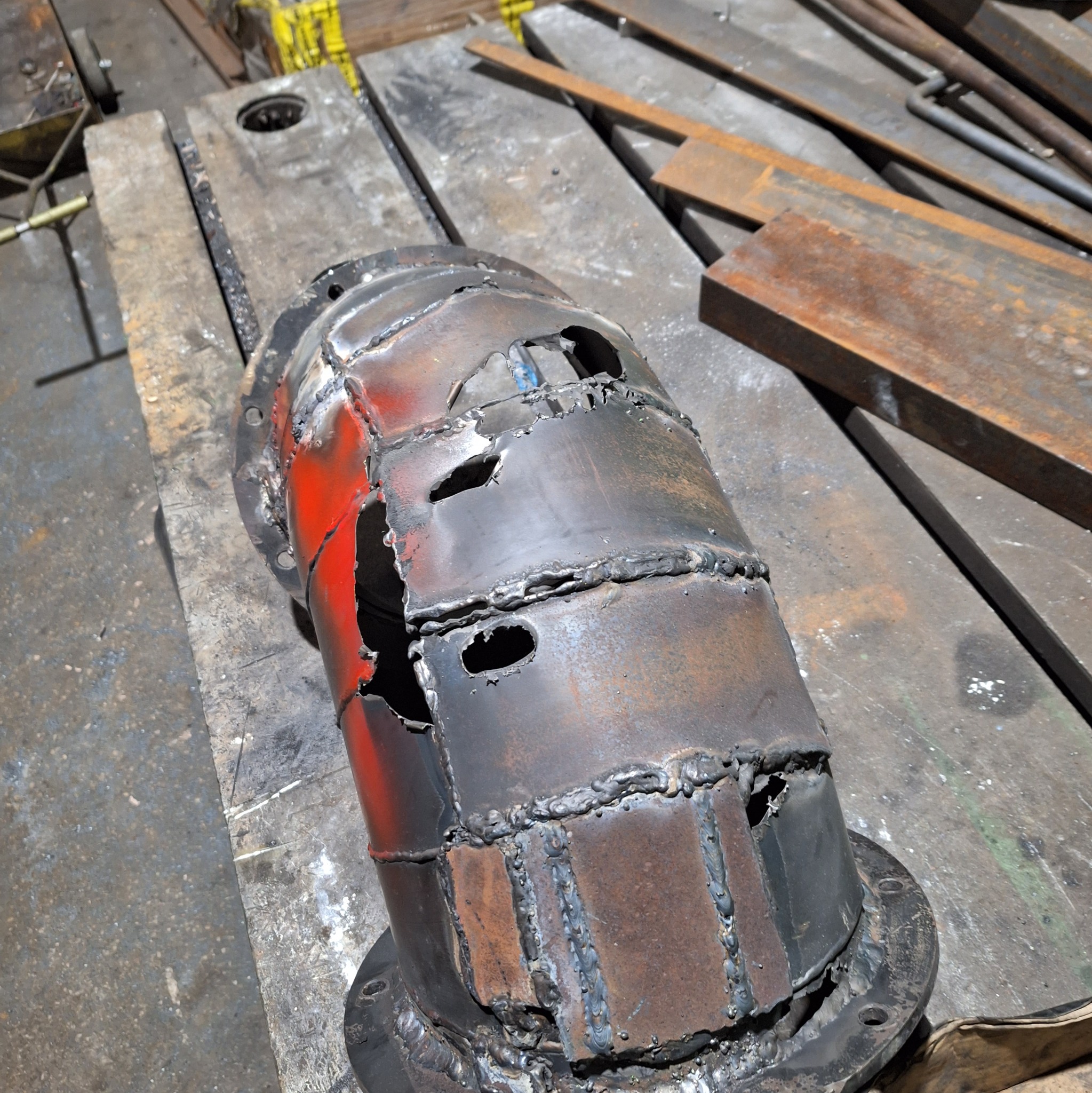 Uszkodzony, spawany metalowy cylinder z otworami i śladami rdzy, częściowo pomalowany na czerwono, leżący na drewnianym podłożu w warsztacie.
