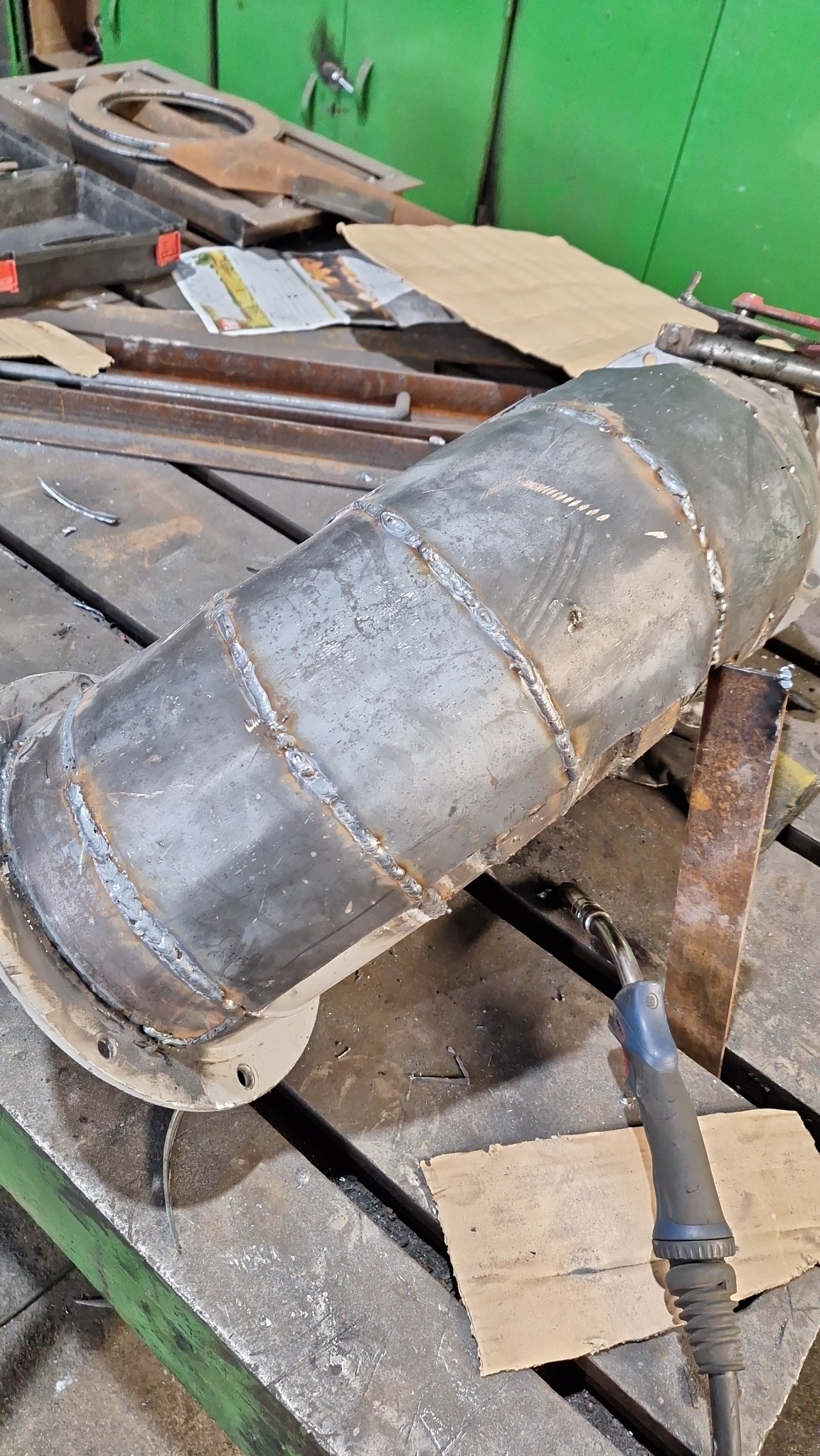 Metalowy cylinder z widocznymi spawami na stole warsztatowym, obok leży palnik spawalniczy, a w tle widać elementy metalowe i zieloną szafkę.
