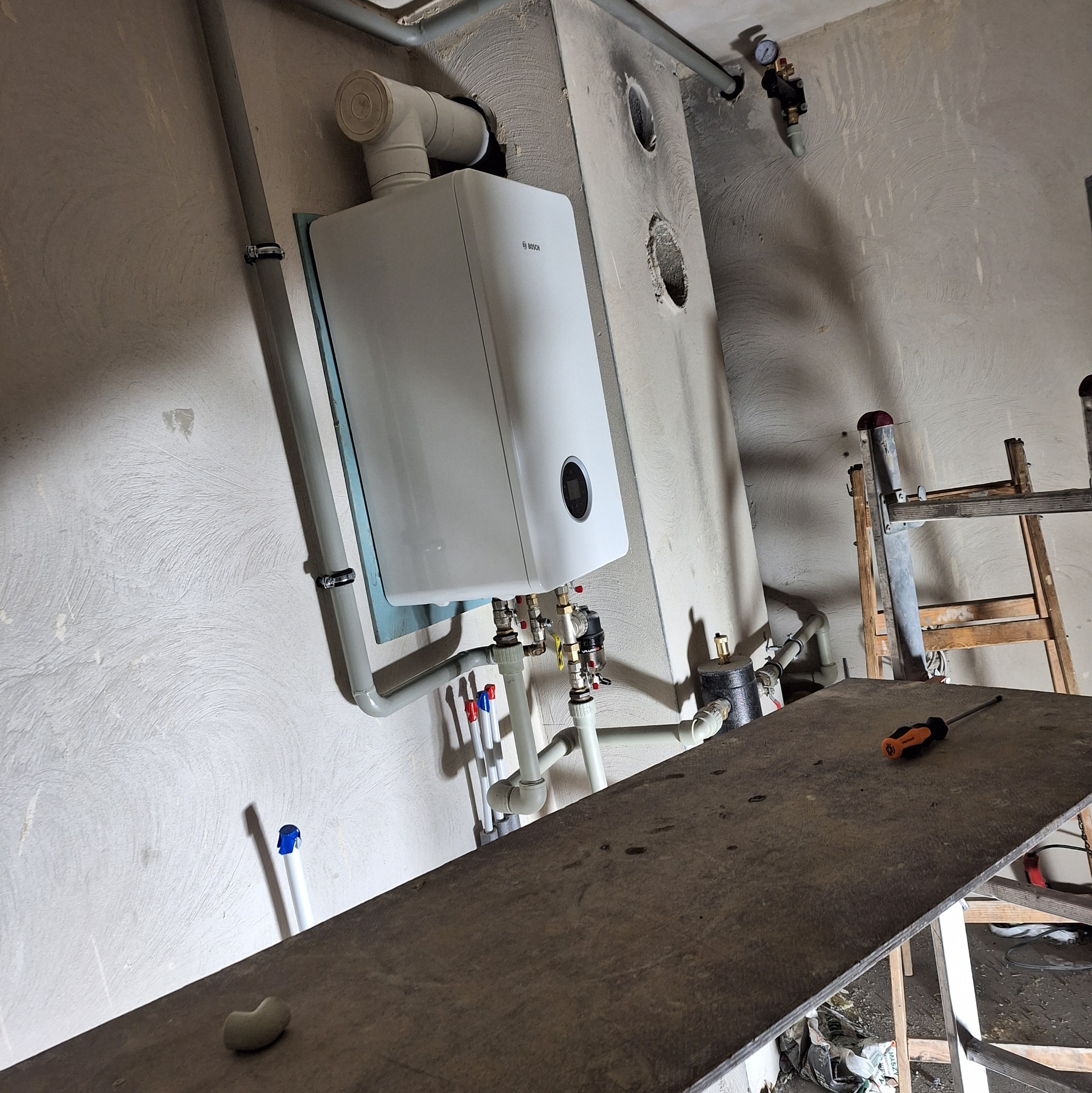 Biały piec gazowy marki Bosch zamontowany na ścianie w trakcie instalacji, widoczne rury, zawory, manometr oraz prowizoryczny stół roboczy ze śrubokrętem i drabina.