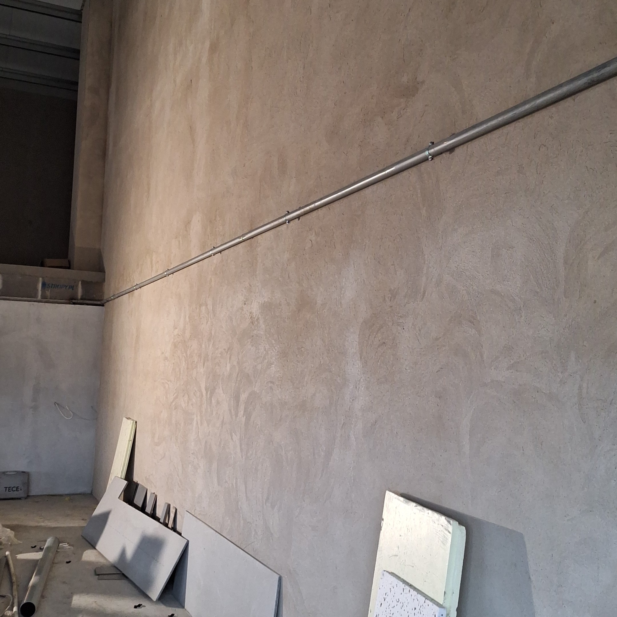 Szara ściana z efektem dekoracyjnym, poziomo poprowadzona metalowa rura przymocowana do ściany, na podłodze płyty gipsowe i styropian.