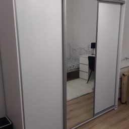 Radosław Monter - Biała szafa przesuwna z lustrem, odbijająca fragment sypialni z łóżkiem i biurkiem, na podłodze panele.