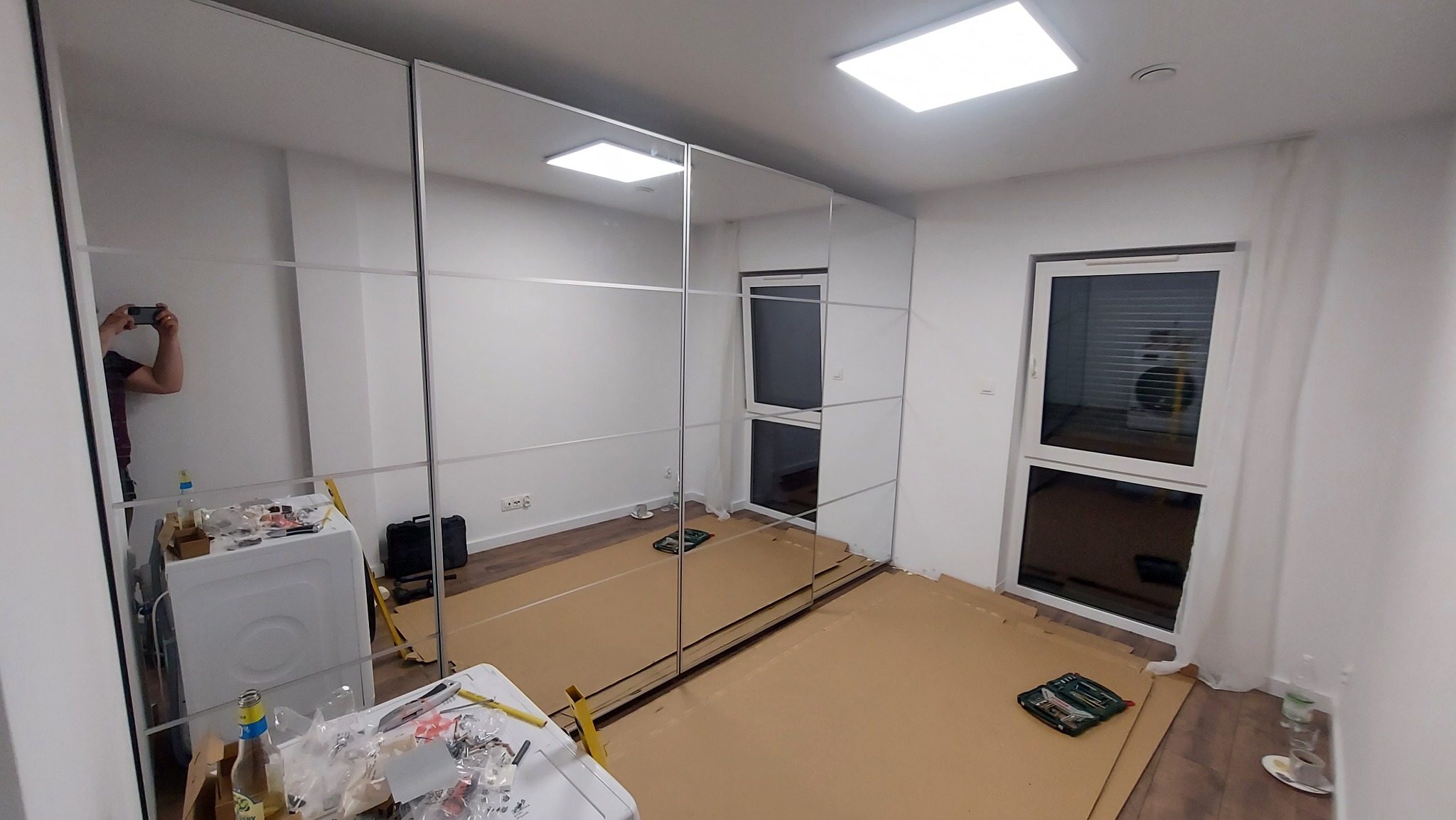 Nowoczesna, przesuwna szafa z lustrzanymi drzwiami w trakcie montażu, zabezpieczona kartonem, z odbiciem osoby robiącej zdjęcie telefonem.