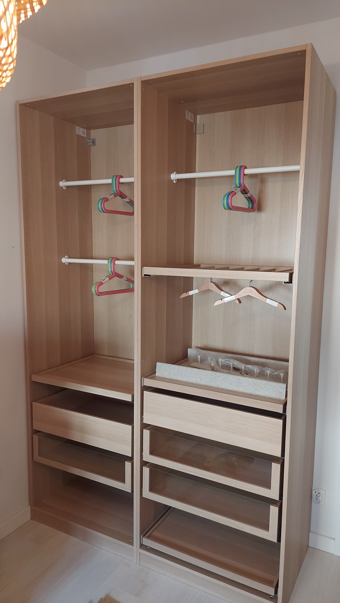 Wnętrze dwuczęściowej, jasnej garderoby z wysuwanymi szufladami i drążkami na wieszaki, zamontowanej we Wrocławiu, z widocznymi wieszakami w kolorowe paski i drewnianymi wieszakami na spodnie.