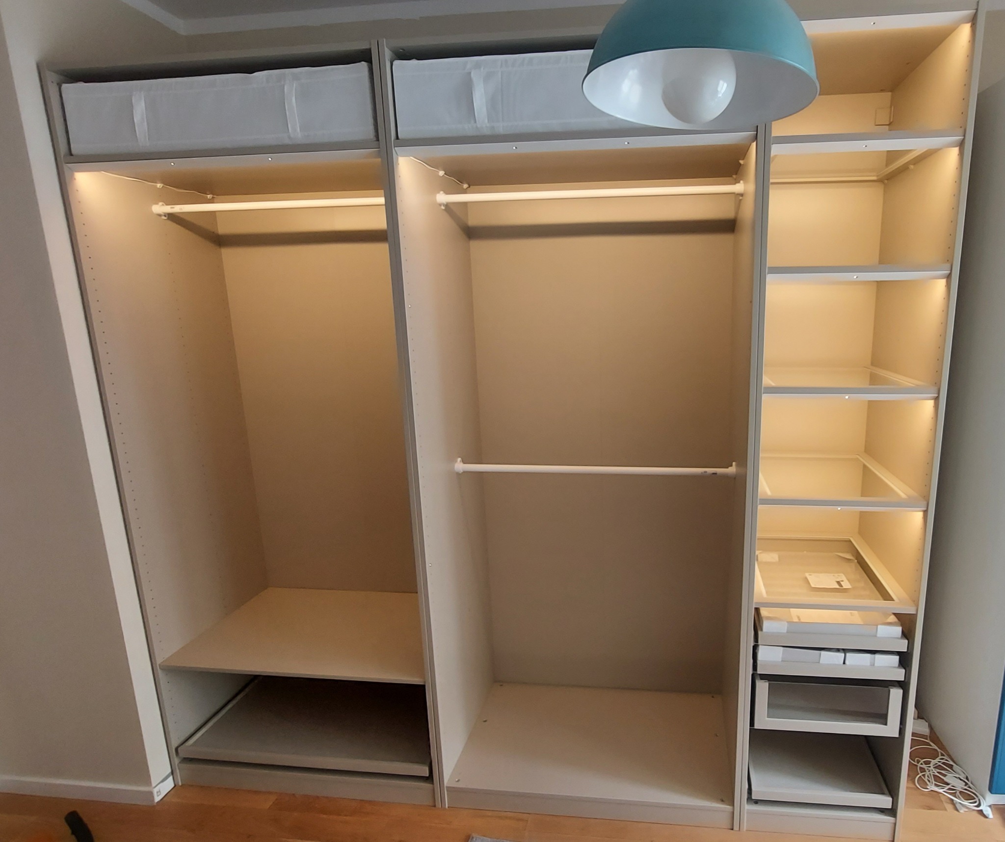Wnętrze szafy modułowej z oświetleniem LED, półkami i drążkiem na ubrania, widoczne białe pojemniki na górze i wysuwane szuflady na dole.