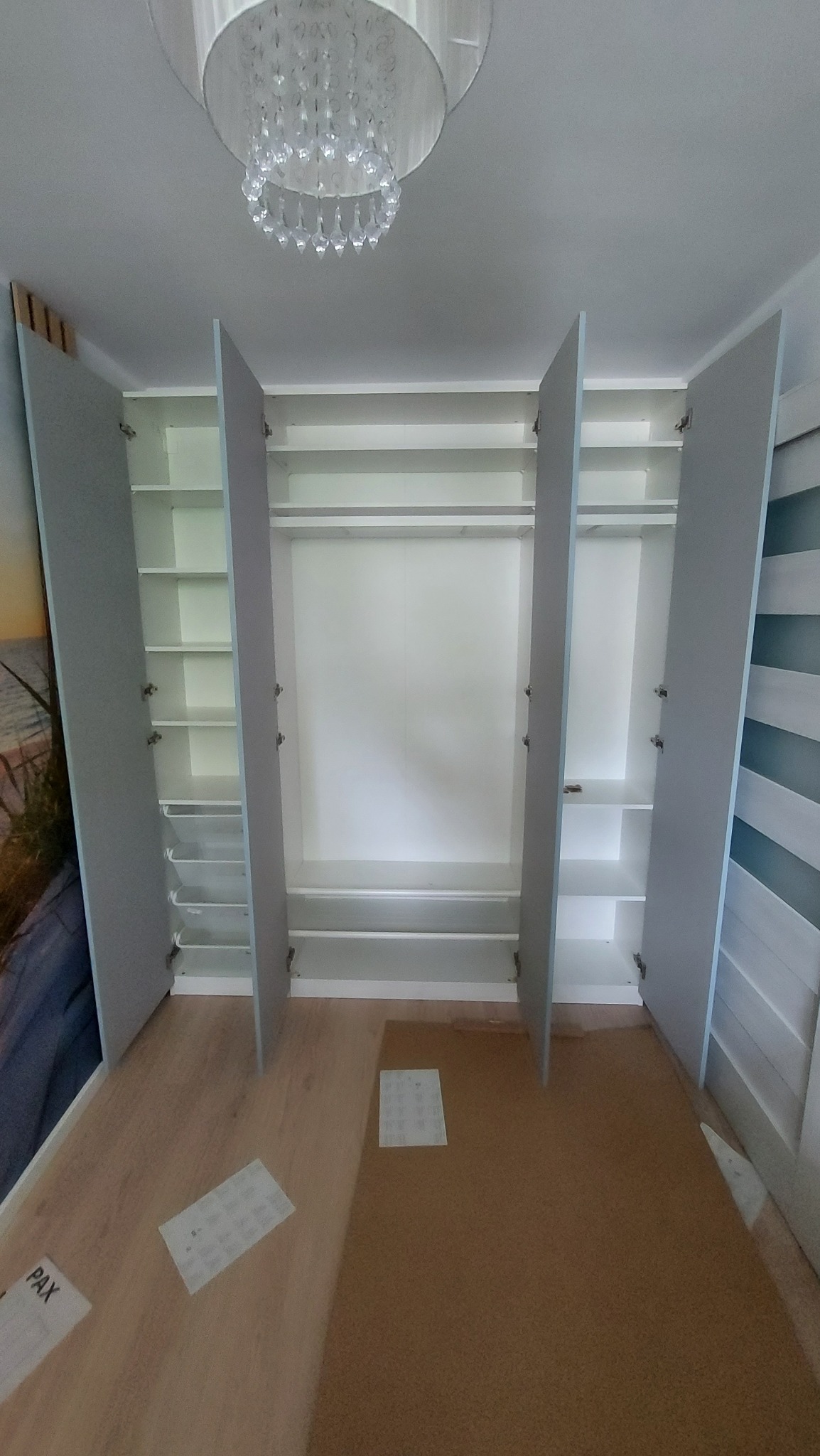 Rozłożona szafa z otwartymi drzwiami, widoczne półki i drążki, na podłodze leżą kartki z instrukcjami, nad szafą wisi kryształowy żyrandol.