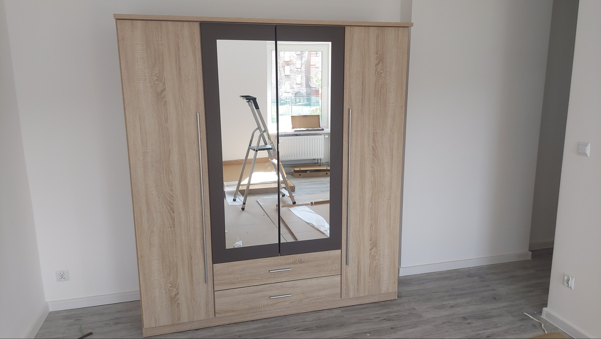 Czterodrzwiowa szafa z lustrem pośrodku, widoczna drabina w odbiciu lustrzanym, kartony na podłodze, jasne panele podłogowe.