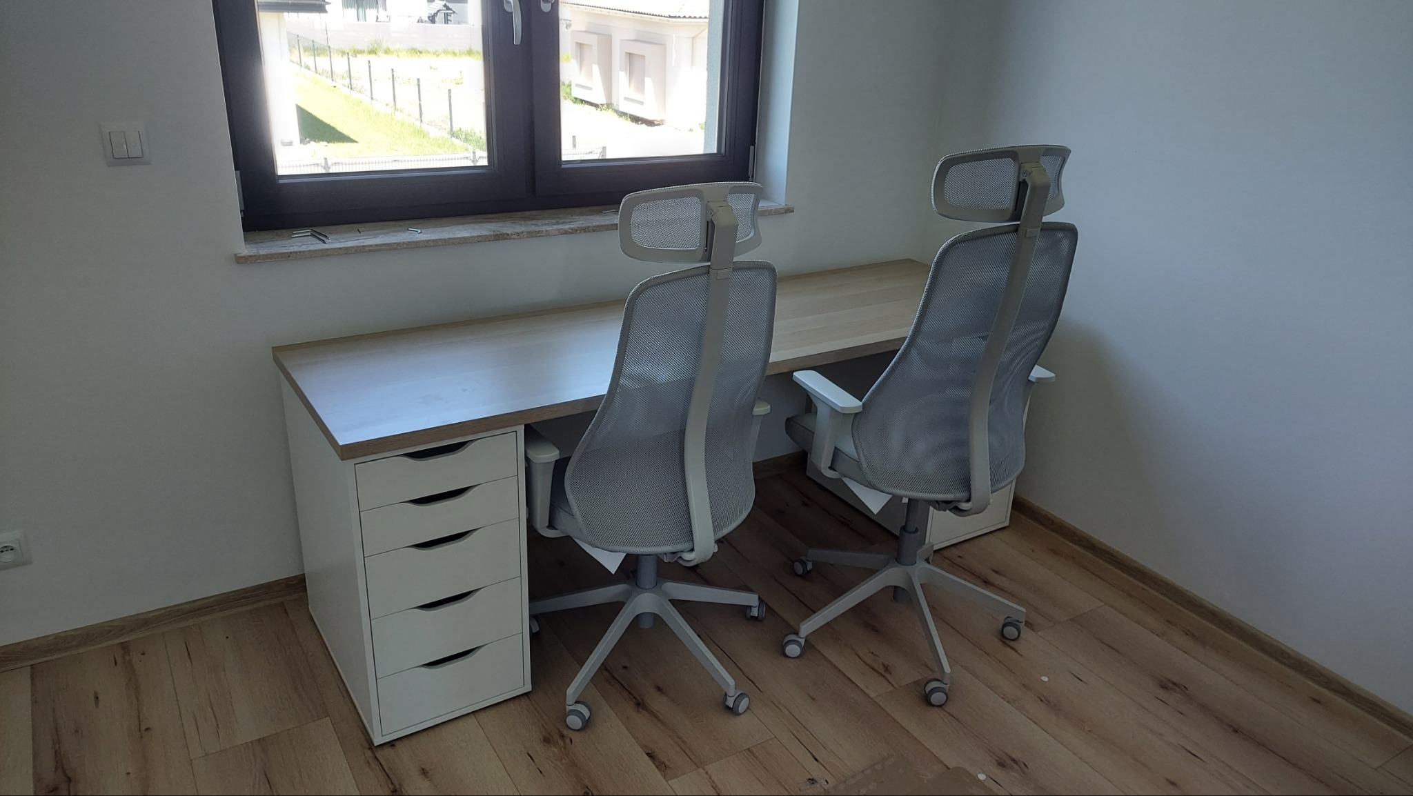 Jasny, minimalistyczny pokój z biurkiem z szufladami i dwoma ergonomicznymi krzesłami biurowymi z siatkowanym oparciem, widok z tyłu na tle okna.