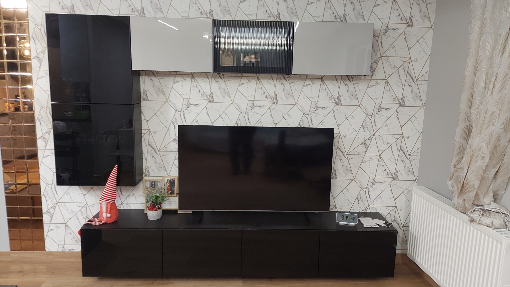 Czarny zestaw mebli RTV z wiszącymi szafkami na tle tapety w geometryczne wzory, z dekoracyjnym krasnalem i ramkami ze zdjęciami na szafce.