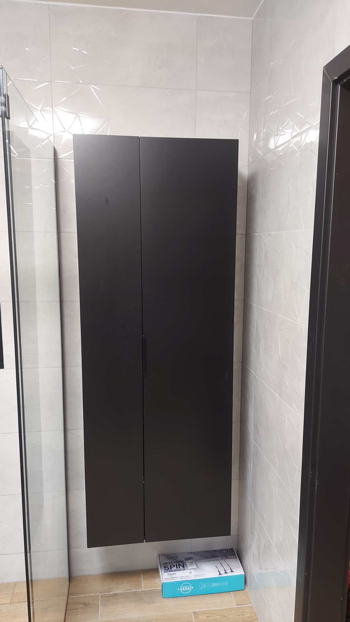 Ciemna szafka łazienkowa zamontowana na ścianie z biało-szarymi płytkami, obok kabiny prysznicowej i drzwi, z pudełkiem nowego sprzętu do czyszczenia pod nią.