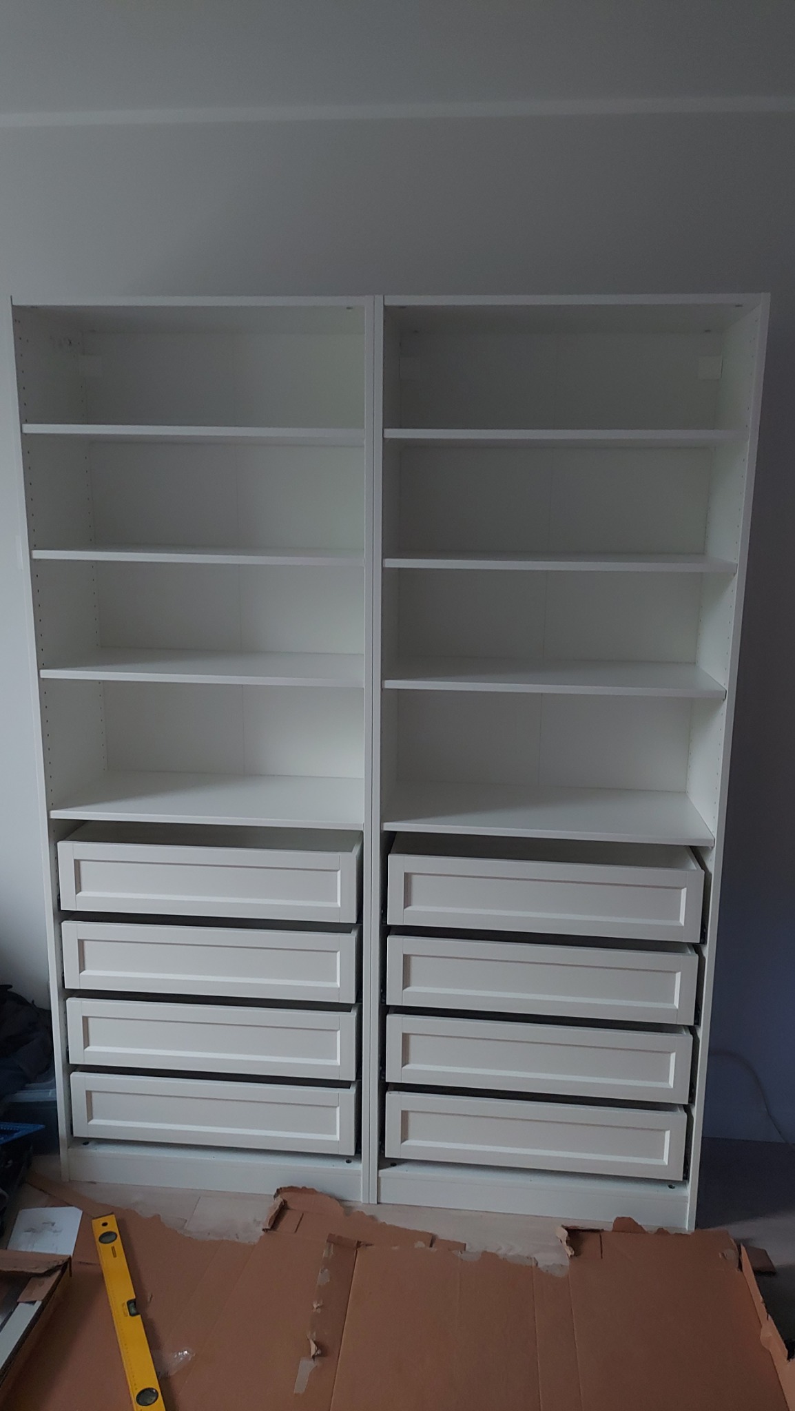 Dwa białe regały z szufladami, częściowo złożone, na podłodze karton i poziomica, montaż mebli w trakcie.