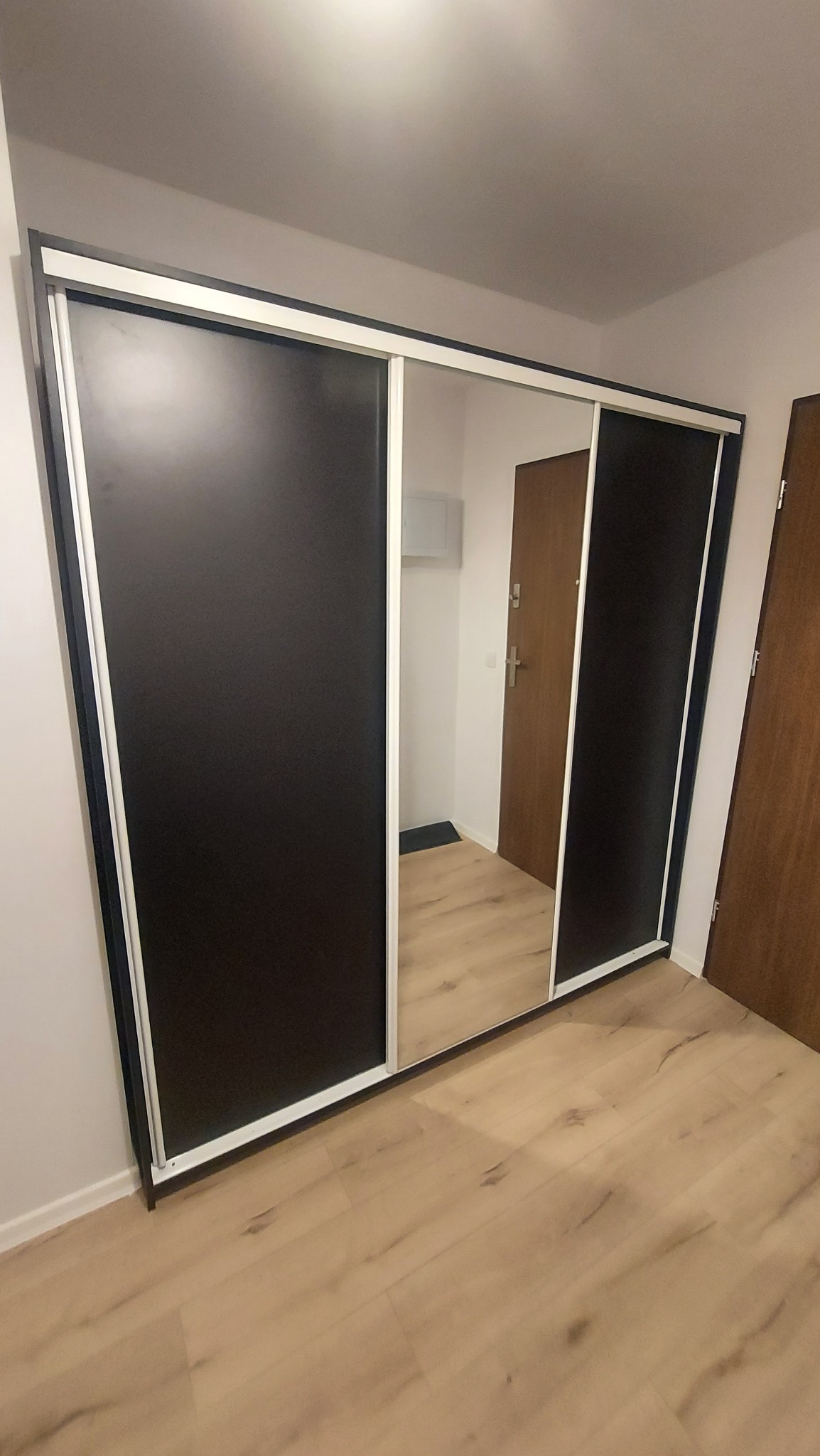 Szafa przesuwna z czarnymi frontami i lustrem, widok na przedpokój z drewnianymi drzwiami.