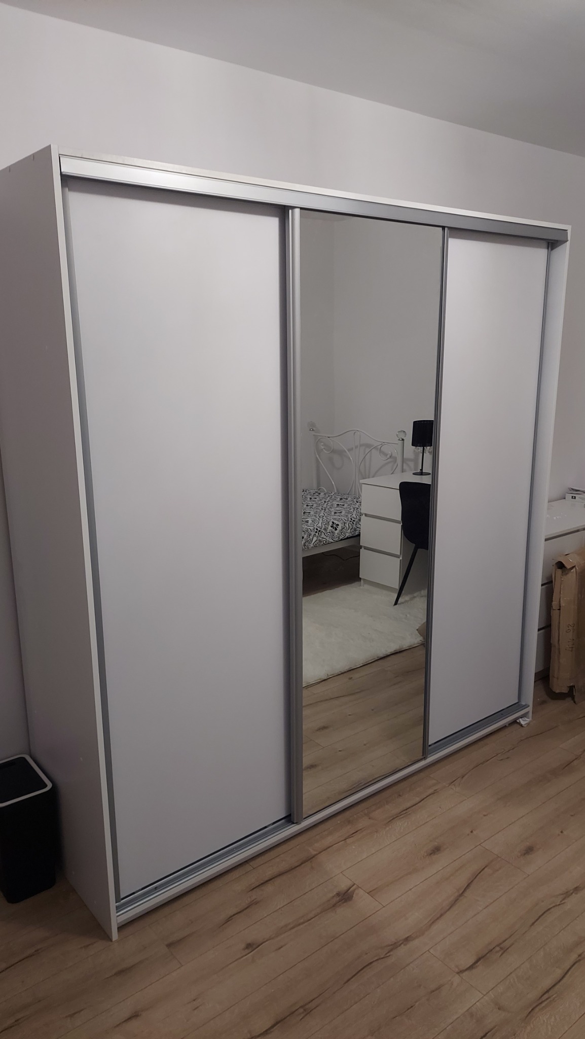 Biała szafa przesuwna z lustrem, odbijająca fragment sypialni z łóżkiem i biurkiem, na podłodze panele.