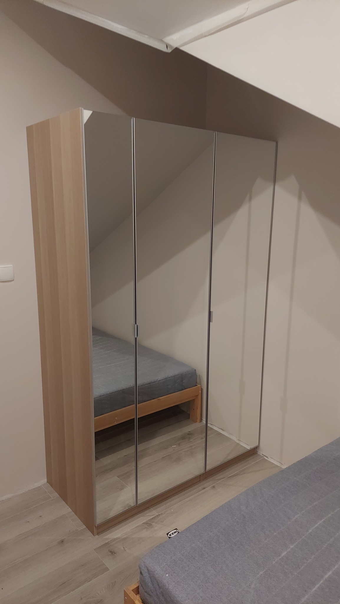 Trzydrzwiowa szafa z lustrami po montażu, odbijająca fragment pokoju z łóżkiem, ustawiona na drewnianej podłodze w pomieszczeniu ze skośnym sufitem.