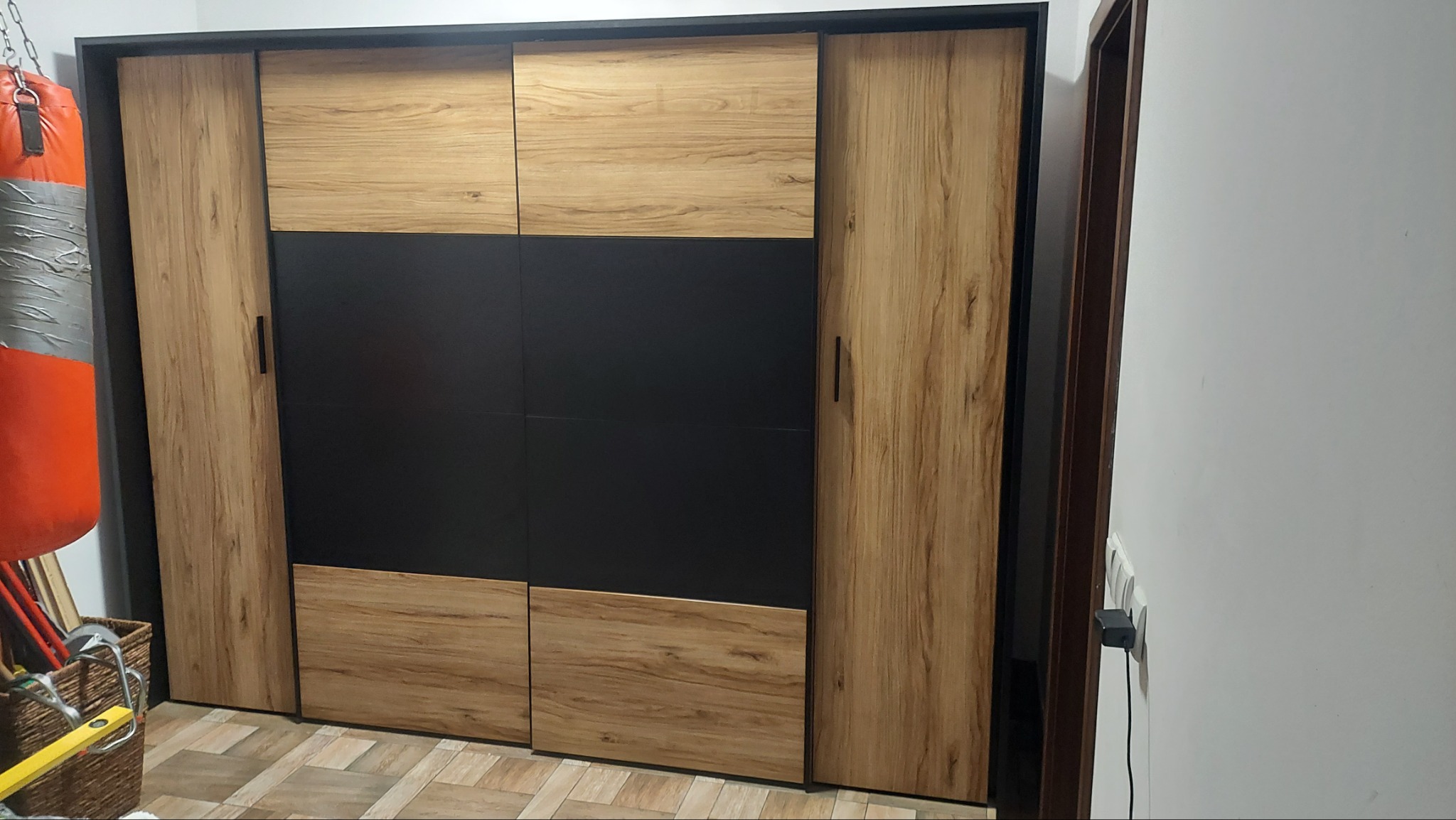 Zamknięta szafa przesuwna z drewnopodobnymi i czarnymi panelami, widoczny worek bokserski w tle, na podłodze panele imitujące drewno.