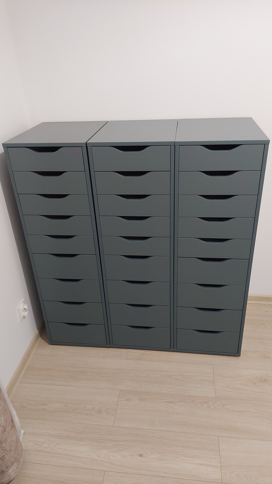 Trzy szare komody z szufladami, ustawione obok siebie, na jasnej podłodze, widoczne gniazdko elektryczne na ścianie.