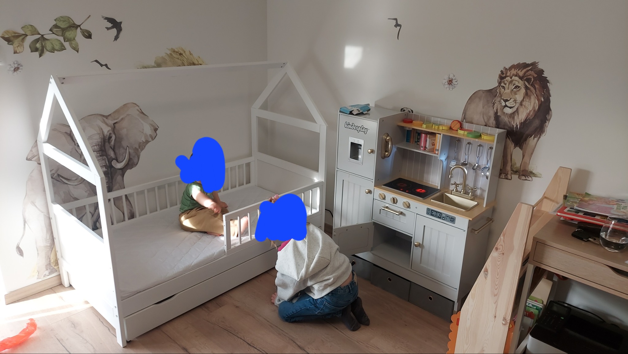 Pokój dziecięcy z białym łóżkiem w kształcie domku, drewnianą kuchnią zabawkową i naklejkami ze zwierzętami na ścianie, w pokoju bawią się dzieci.