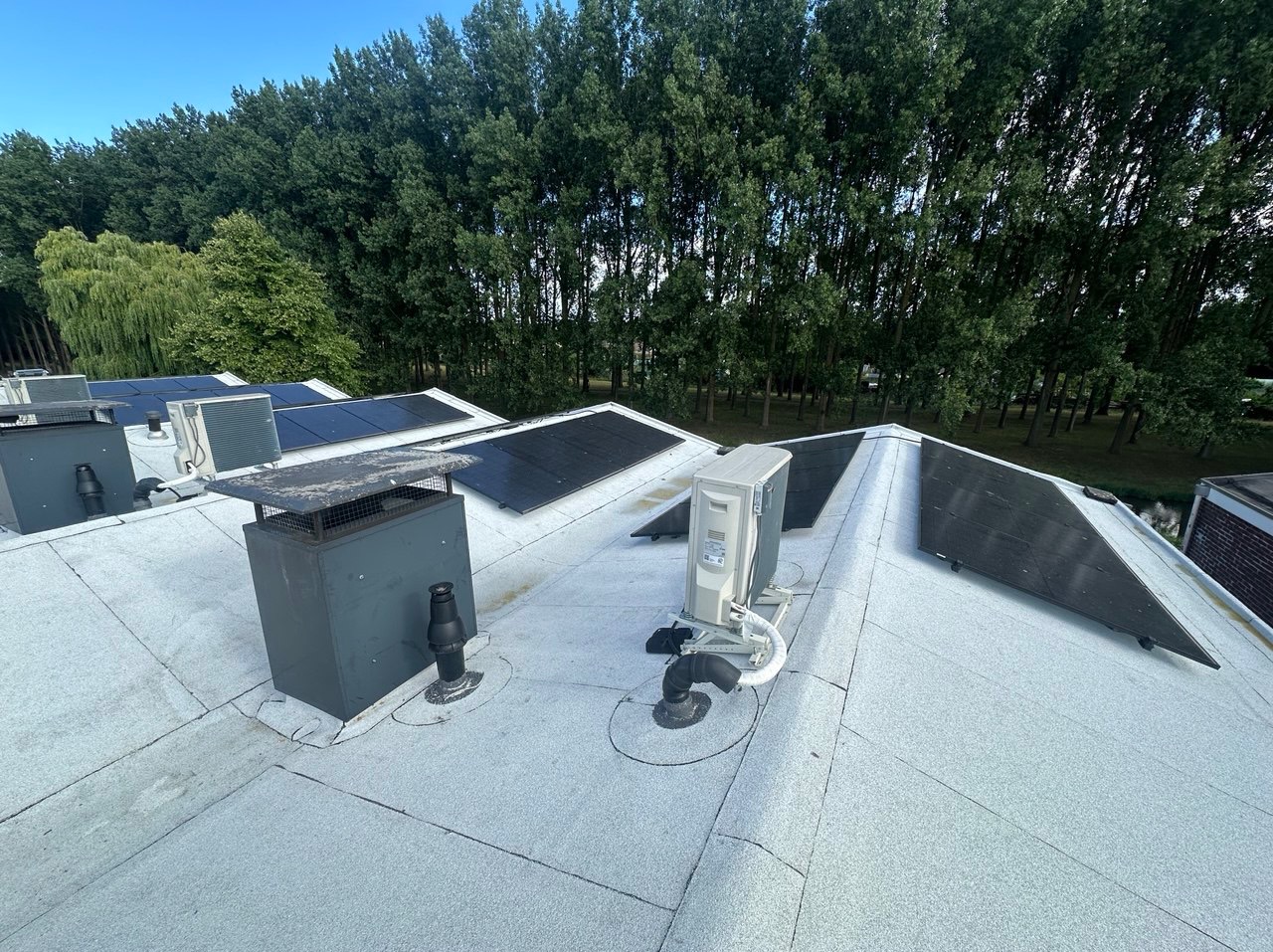 Biały dach pokryty papą z panelami słonecznymi i klimatyzatorem na tle zielonych drzew. Widok z góry, oświetlenie dzienne.