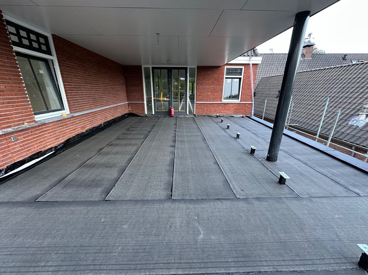 Płaski dach pokryty czarną papą, widoczne łączenia i elementy mocujące. Czerwona ceglana ściana budynku w tle, okna z białymi ramami. Na dachu stoi gaśnica.