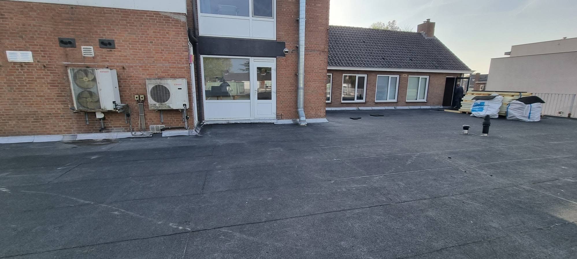 Wykończony dach płaski z papy termozgrzewalnej na budynku z cegły, widoczne kominy wentylacyjne, klimatyzatory na ścianie i materiały budowlane przygotowane do dalszych prac.