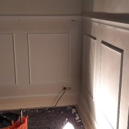 Leszek Zapolski - Ściana z zamontowaną dekoracyjną sztukaterią w postaci paneli ściennych, widoczne gniazdko elektryczne i narzędzia na podłodze.