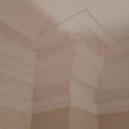 Leszek Zapolski - Wykończony narożnik sufitu z listwą dekoracyjną, świeżo pomalowany na biało, widoczne delikatne niedoskonałości na ścianie w kolorze beżowym.