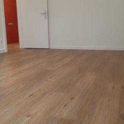 Leszek Zapolski - Jasne panele podłogowe imitujące drewno w pustym pokoju z białymi ścianami i pomarańczowymi drzwiami w tle.