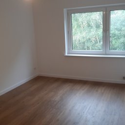 Leszek Zapolski - Pusty pokój po remoncie z drewnianą podłogą, białymi ścianami i oknem z widokiem na zieleń, listwy przypodłogowe pomalowane na biało, widoczne gniazdka elektryczne.