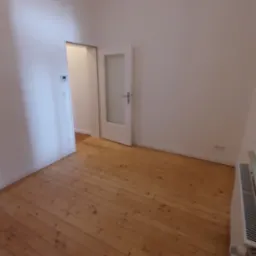 Pusty pokój z drewnianą podłogą, białymi ścianami, grzejnikiem i otwartymi białymi drzwiami z szybą. Widoczny fragment kolejnego pomieszczenia.