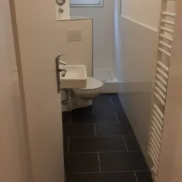 Wykończona łazienka w Bojanowie z ciemną, prostokątną glazurą podłogową, białą armaturą (umywalka i toaleta) oraz grzejnikiem drabinkowym. Widoczny fragment kabiny prysznicowej.