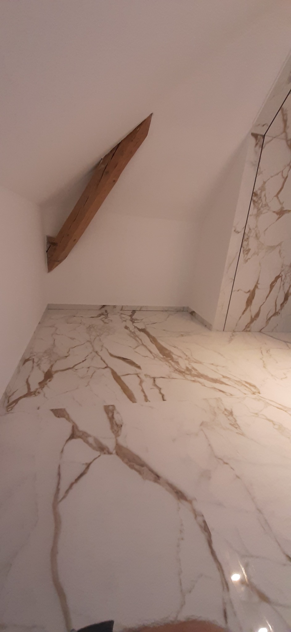 Wykończone wnętrze poddasza ze skośnym sufitem, z drewnianą belką konstrukcyjną i białymi ścianami. Na podłodze i fragmencie ściany płytki imitujące marmur z wyraźnym, brązowym żyłowaniem.