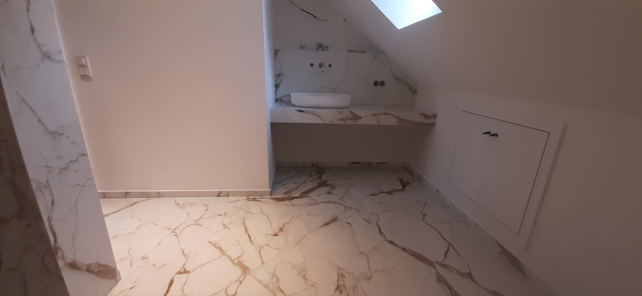 Wykończona łazienka na poddaszu ze skośnym sufitem i oknem dachowym, białe płytki imitujące marmur z brązowymi żyłami na podłodze i ścianie za umywalką, biała umywalka nablatowa, białe ściany...