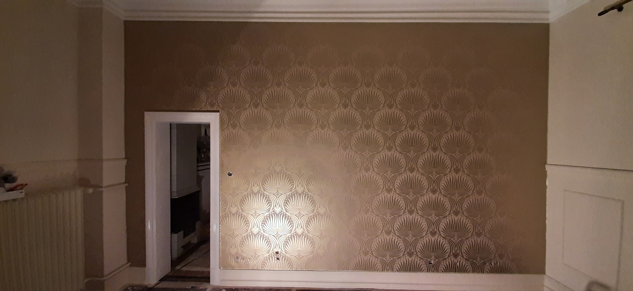 Ściana w pokoju pokryta tapetą w odcieniach złota z wzorem wachlarzy, widoczne ślady po niedokładnym przyklejeniu w dolnej części, biała listwa przypodłogowa i framuga drzwi po lewej stronie.