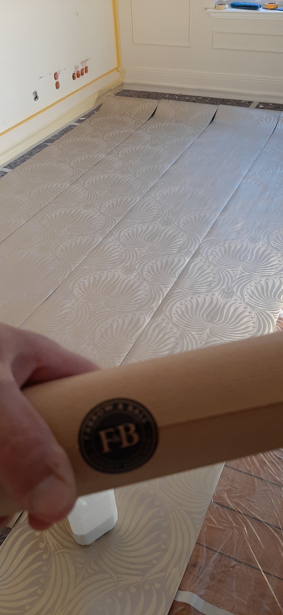Przygotowane pasy tapety z delikatnym wzorem ułożone na podłodze, widoczna naklejka z logo 'FB' na opakowaniu, zabezpieczona taśmą ściana w tle.