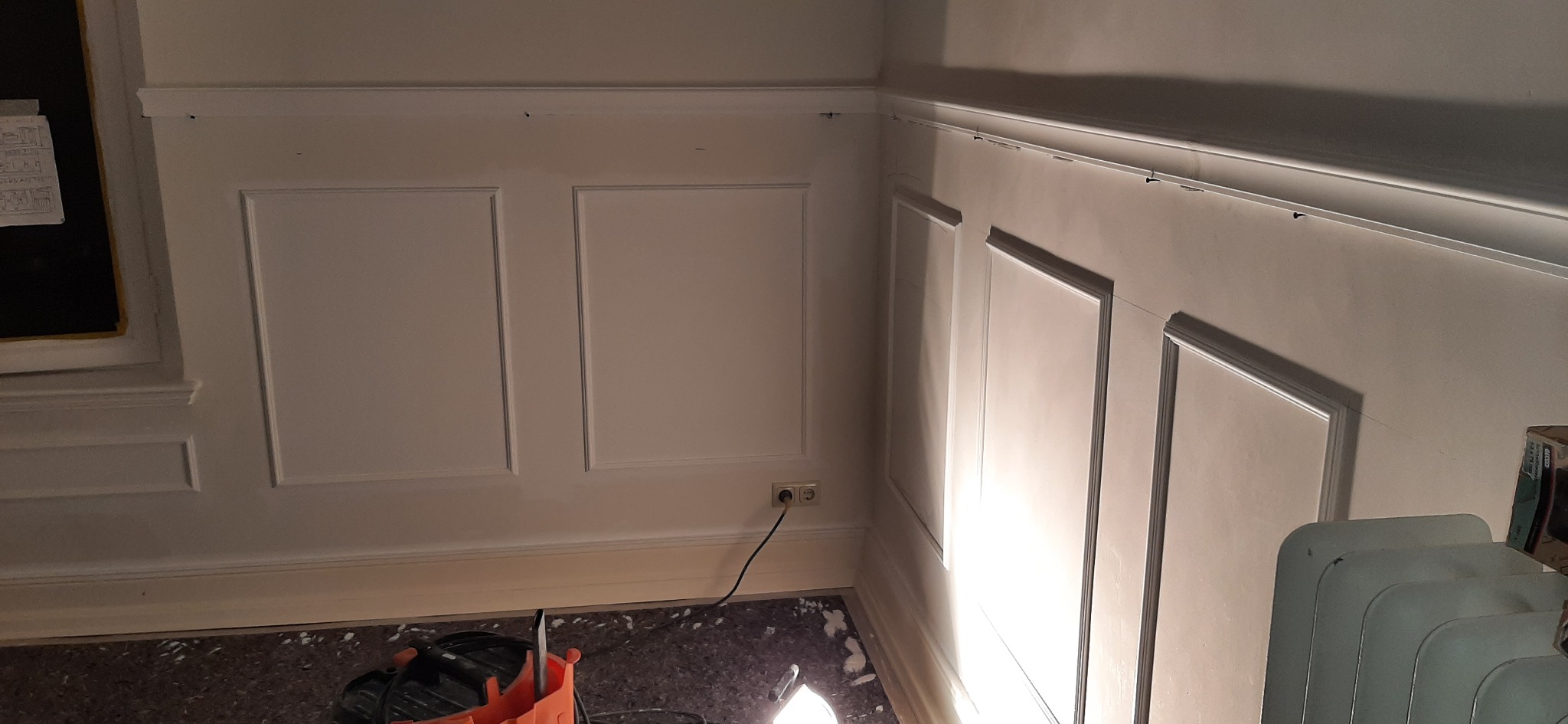 Ściana z zamontowaną dekoracyjną sztukaterią w postaci paneli ściennych, widoczne gniazdko elektryczne i narzędzia na podłodze.